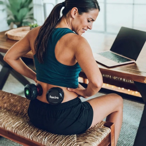Woman using a Fasica muscle stimulator at a desk with a laptop.